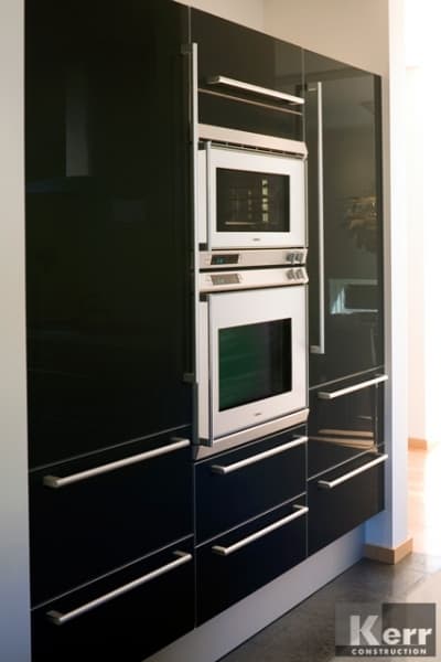 Sleek modern kitchen renovation with glossy black cabinets featuring built-in stainless steel microwave and oven, vertical handles, and reflective surfaces enhancing the contemporary design. Kerr Construction logo in the corner.