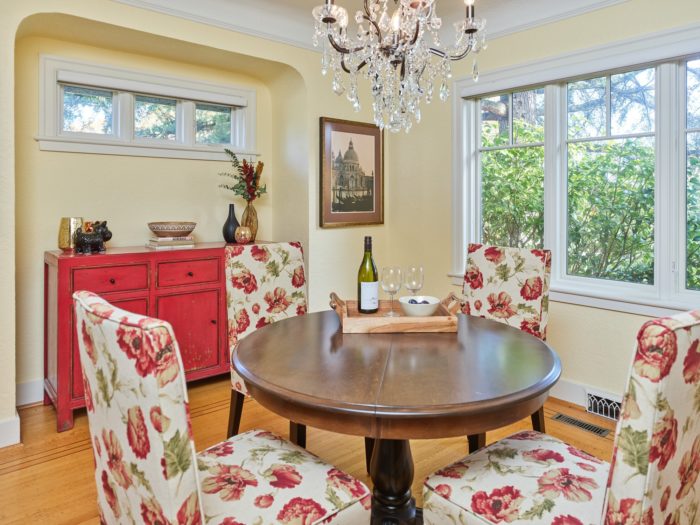 A dining room transformation from drab to fab features a round wooden table surrounded by four chairs with floral cushions. A chandelier glimmers above, while a red sideboard displays decor. A large window showcases lush trees, and wine glasses sit elegantly beside a framed picture on the wall.