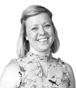 A woman with shoulder-length hair, wearing a floral-patterned, sleeveless top, exudes the warmth and charm youd expect from home design experts. She smiles confidently at the camera against a plain white background.