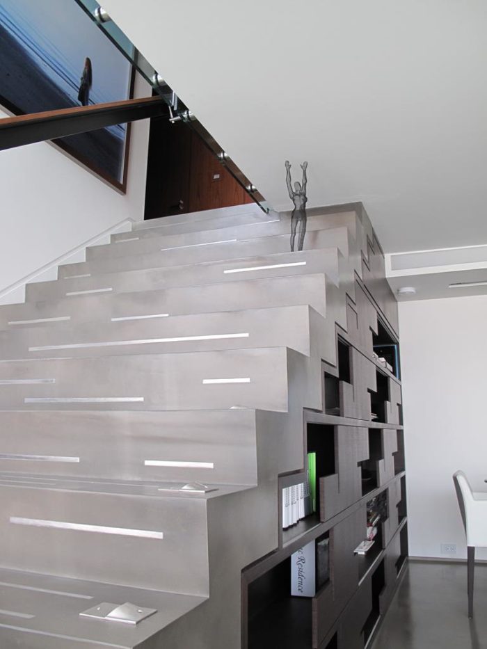 Modern staircase design featuring a sleek, angular style with dark-brown shelving integrated below. A small abstract sculpture graces a step, while a large framed photograph adorns the wall above. The space is well-lit and minimalist, epitomizing sophisticated simplicity.