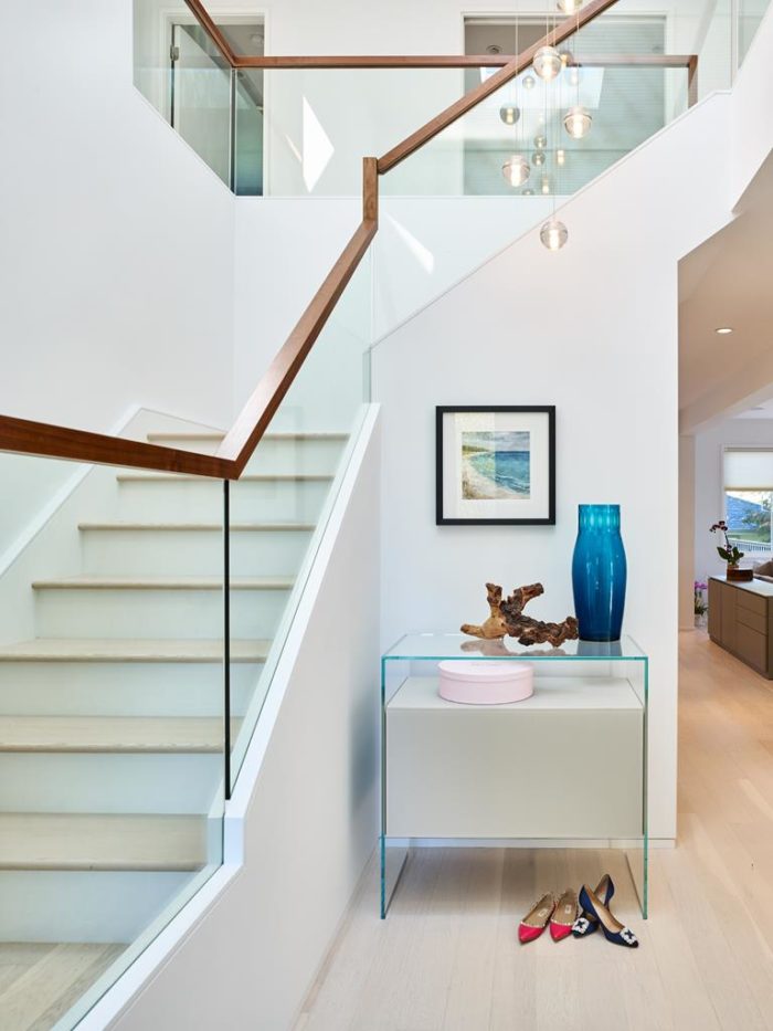 A modern staircase design features glass panels and wooden handrails. Below, a glass console holds a blue vase, a wood sculpture, and two pairs of shoes. A framed artwork hangs on the wall above while pendant lights elegantly dangle from the ceiling.