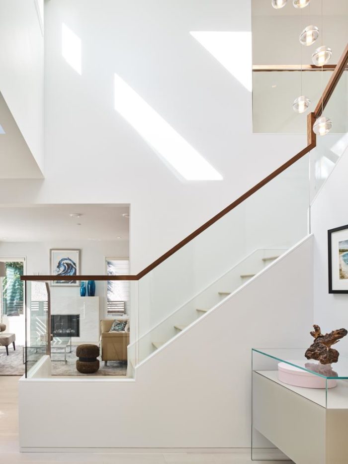 A modern staircase design takes center stage in this white-walled interior, featuring glass railings and wooden handrails. Hanging lights illuminate the space, where sunlight streaming in highlights the artwork, fireplace, and a sculptural piece on a glass table.