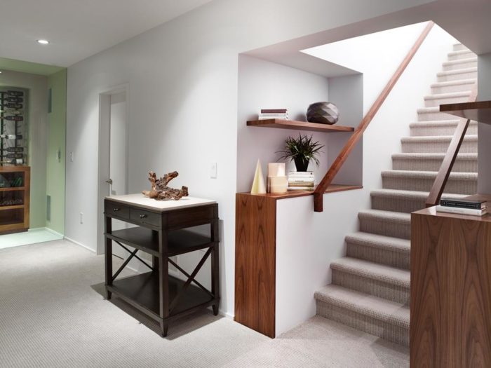 A modern staircase design showcases a carpeted staircase with a sleek wooden handrail. Wooden shelves above display a plant and decorative items. To the left, a small console table holds books and sculpture. The white walls contribute to the bright, airy atmosphere.