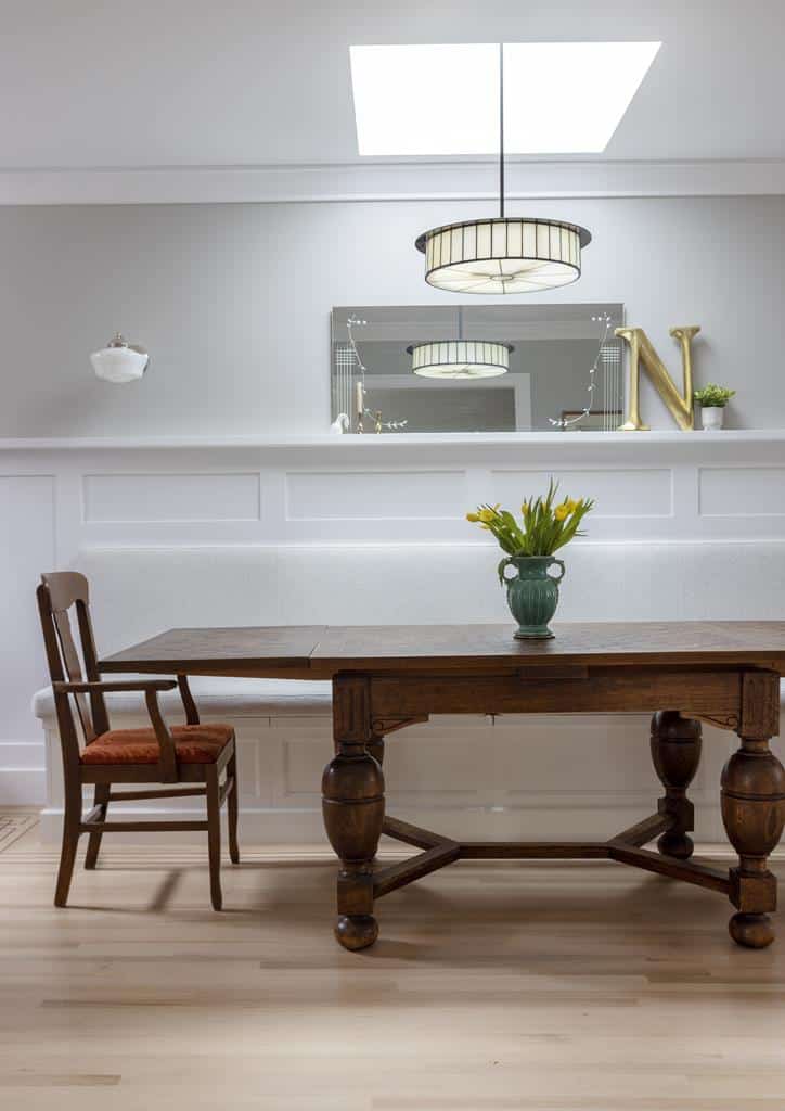 A wooden dining table with ornate legs graces the light floor of this Heritage Home. A small vase of yellow flowers adds charm, while a mirror, decorative letter N, and lamp complete the look. A single chair with a red cushion invites relaxation in this stunning interior makeover.