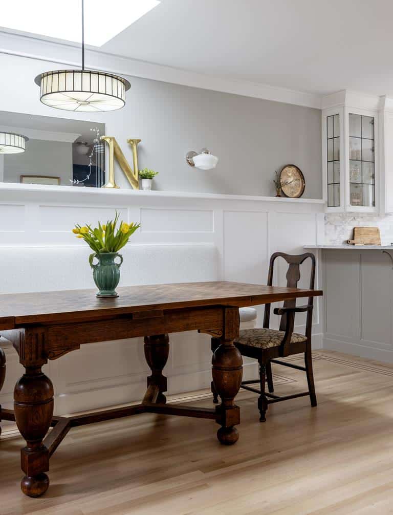 A cozy kitchen in a heritage home features a wooden table and single chair. A vase with yellow tulips graces the table, while gray and white walls host a decorative mirror and clock. A lamp hangs above, complementing the kitchen cabinetry in this charming interior makeover.