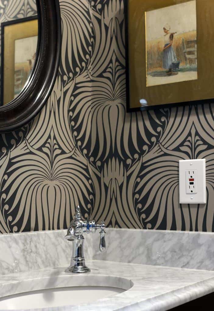 This Heritage Home bathroom exudes elegance with its ornate gray and black wallpaper, a round mirror, and framed art. The marble countertop complements the silver faucet, while a white electrical outlet adds a modern touch to this tasteful interior makeover.