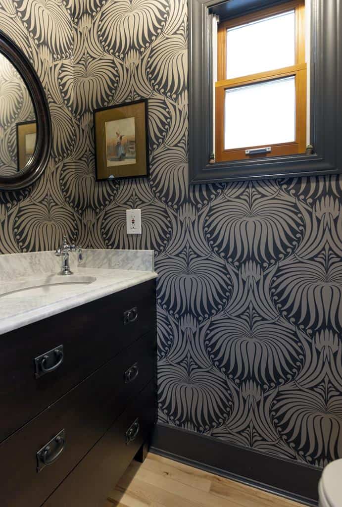 This heritage home bathroom boasts an interior makeover, showcasing black and white ornate patterned wallpaper. It features a black sink cabinet with a marble countertop, a round mirror, and a small window with wooden trim. A framed picture adds charm to the wall.