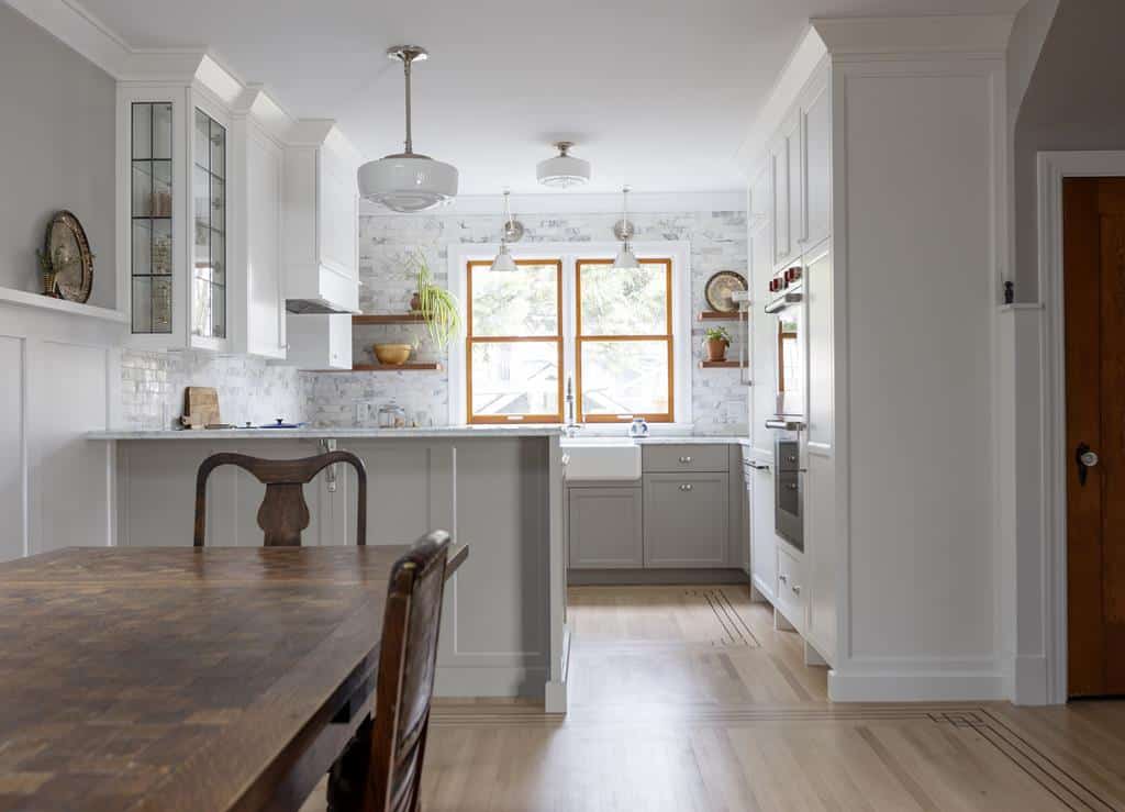 A bright, modern kitchen in a heritage home features white cabinets, a farmhouse sink, and a wooden table. Large windows flood the space with natural light. Two pendant lights hang above the island, and open shelving showcases decor items in this stunning interior makeover.