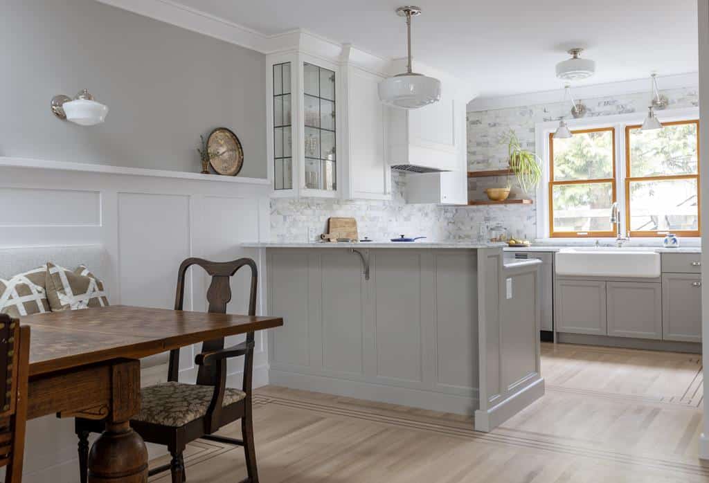 A modern kitchen in a Heritage Home boasts a gray and white color scheme. It features a wooden dining table with chairs, pendant lights, marble backsplash, farmhouse sink, and large window. Light wooden flooring and white paneled walls complete the interior makeover charm.