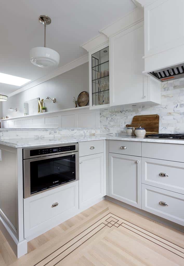 A modern kitchen in a heritage home, featuring white cabinets and marble countertops. It boasts a built-in microwave, cooktop, wooden cutting board, and a small jar on the counter. The light-colored backsplash complements the geometric floor pattern, all illuminated by a pendant light above.