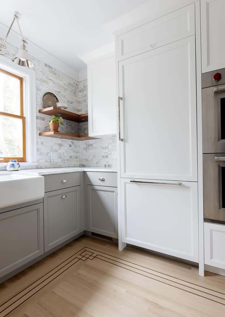 A modern kitchen corner in a charming heritage home features a farmhouse sink beneath a window, light gray cabinets, and a tall white refrigerator. An interior makeover includes shelves with decorative items on the left, while the wooden floor boasts a geometric inlay pattern.