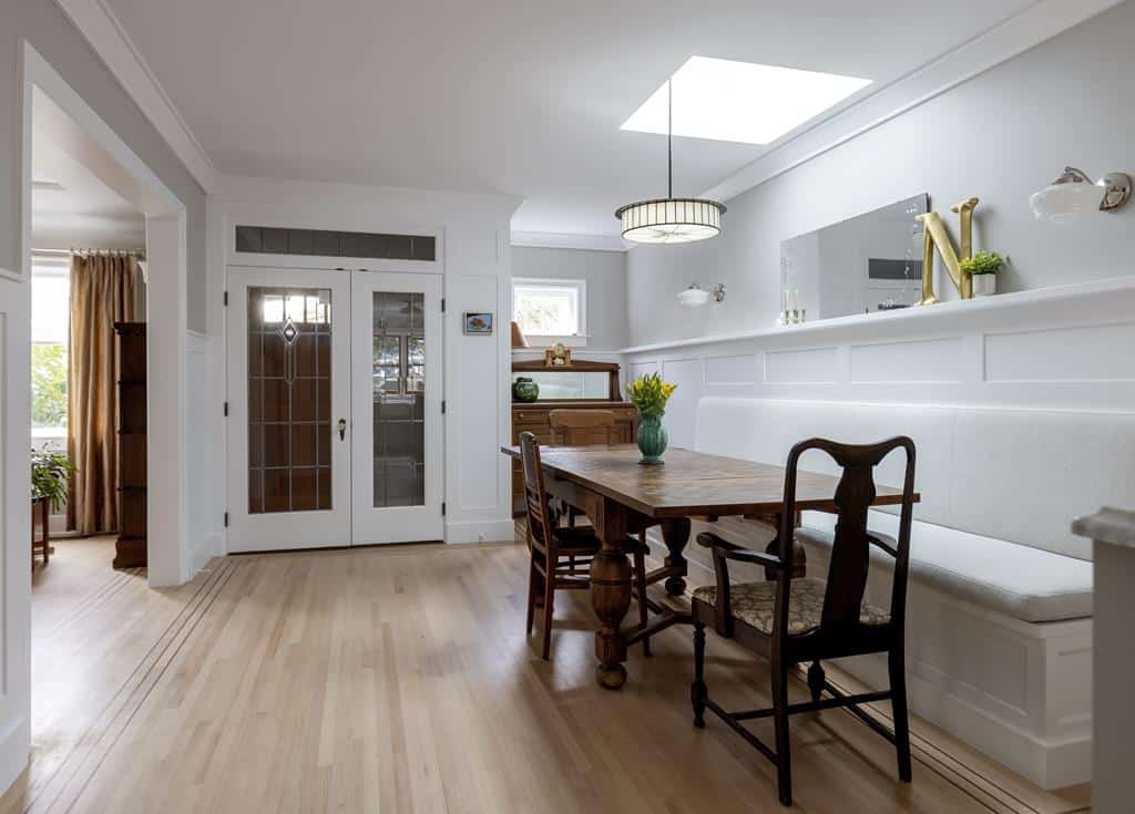 A dining room in this heritage home features a wooden table and assorted chairs on a light wood floor. A built-in bench with cushions lines the wall. Above, a large, circular ceiling light adds charm, while a mirror and small shelf adorn the walls. Doorways open to other rooms, hinting at an inviting interior makeover.