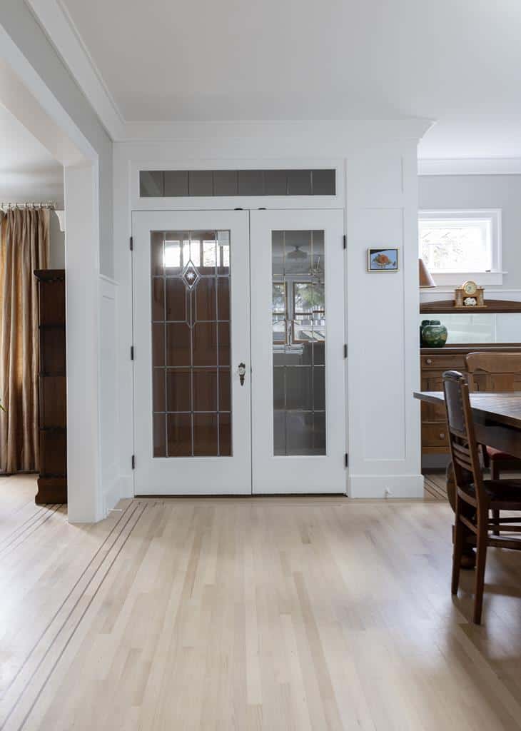 Antique double doors with decorative glass panels are set in a white wall, leading to another room in this heritage home. The surrounding decor includes a wooden cabinet on the left and a dining table with chairs on the right, accentuating the light wooden floorings charm and showcasing an interior makeover.