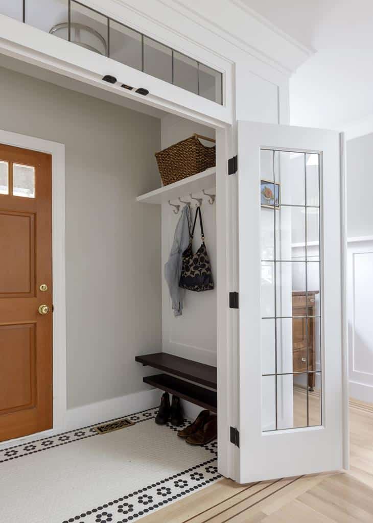 A small entryway in this heritage home features a brown door, a bench, and a shelf above. The shelf holds a woven basket, while hooks beneath hold a black bag and gray scarf. Shoes are neatly placed on the floor beneath the bench. A white folding door is partially open.