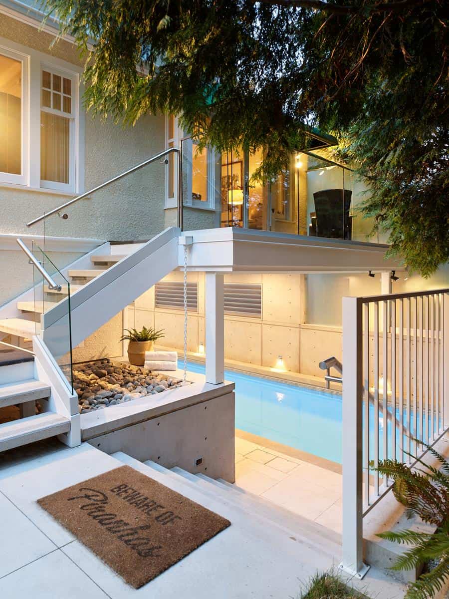 This modern backyard, a contender for renovation awards, features a small, narrow pool tucked below a staircase. Glass railings and warm indoor lighting create an inviting glow through the windows. A welcome mat on the deck reads Beware of Paradise, while lush greenery surrounds this haven.