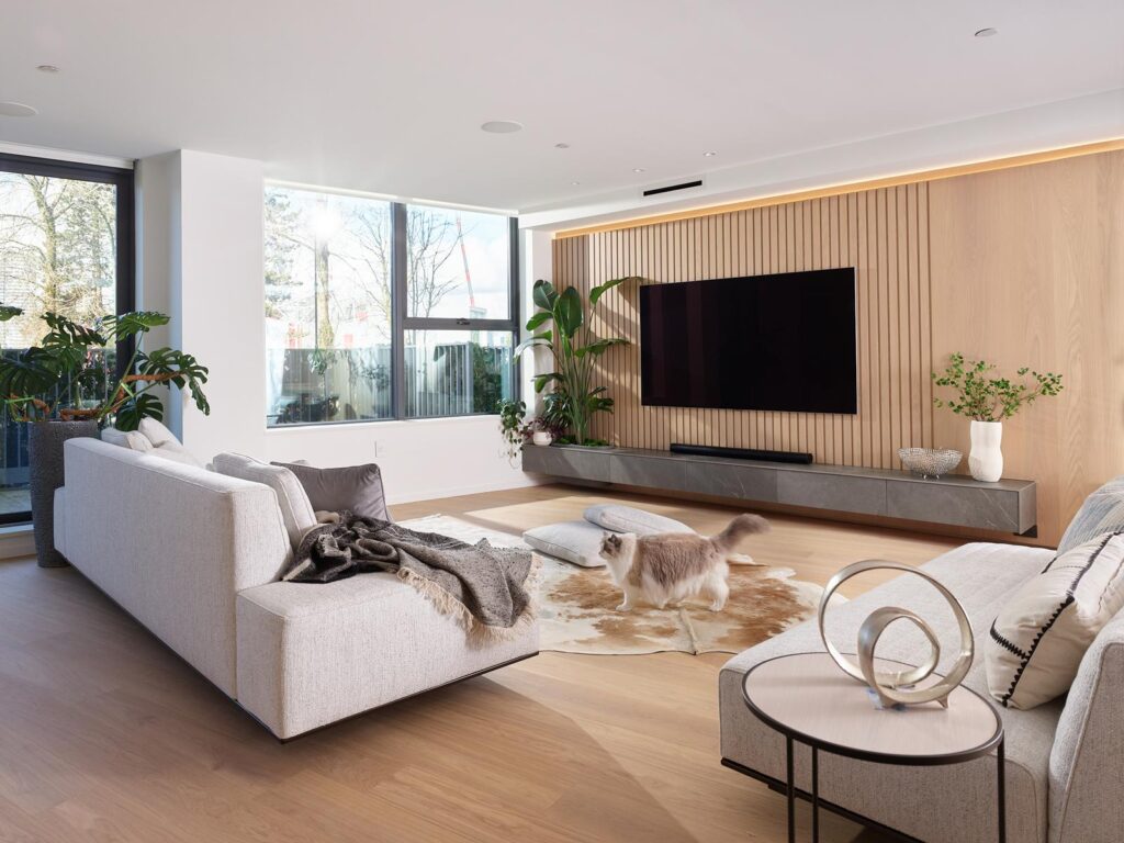Nestled in the heart of Quebec St. Condo, this modern living room boasts light wood floors and a white sofa atop a chic cowhide rug. A cat frolics playfully as green plants by the windows breathe life into this elegant living space, all centered around a sleek TV on a slatted wood accent wall.