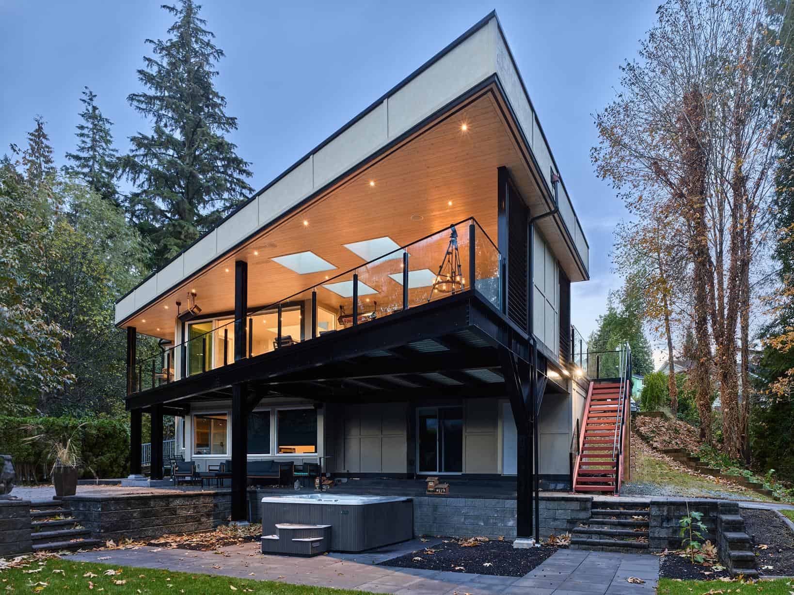 Modern two-story house with large windows, a spacious balcony, and an angled roof, surrounded by trees. Outdoor lighting glows warmly, and a hot tub sits on a beautiful deck in the landscaped yard with a beautiful view.