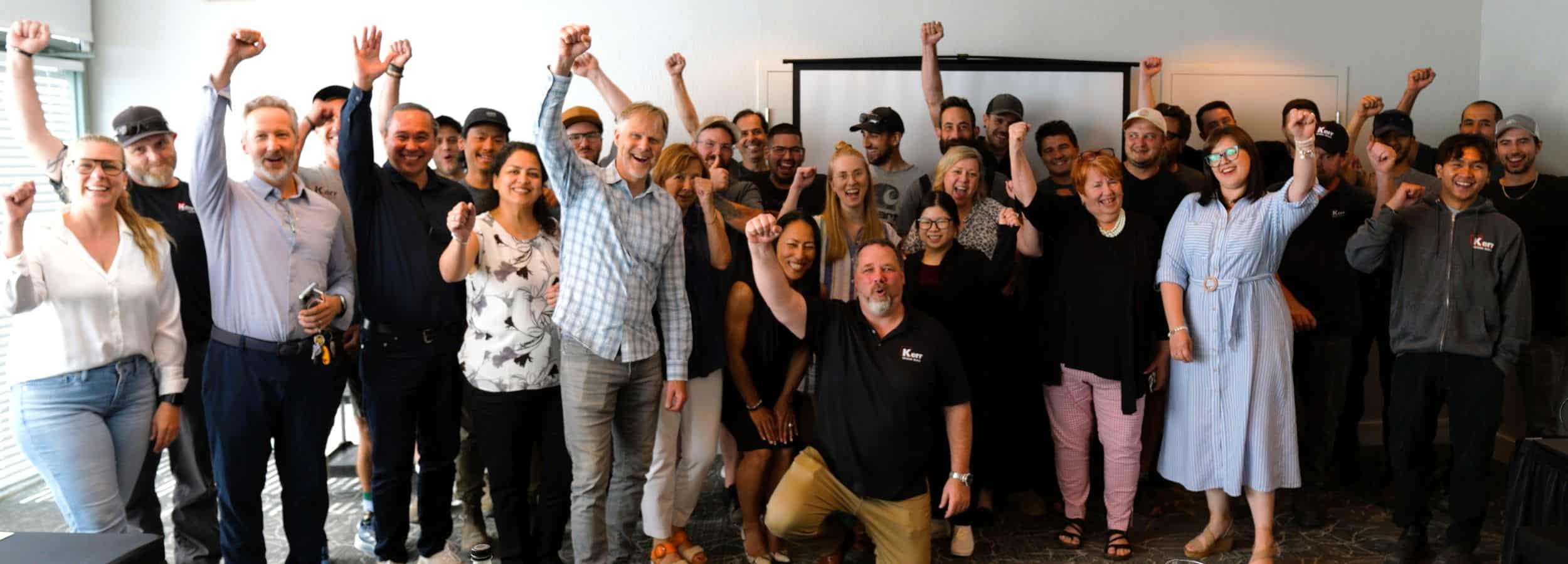 A diverse group of people stand together indoors, smiling and raising their fists in celebration or solidarity—a new era of teamwork and innovation at Kerr Design Build. Most are in casual or business-casual attire, facing the camera enthusiastically.