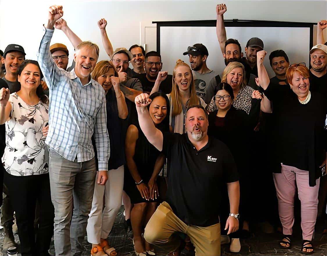 A diverse group of people pose together indoors, many smiling and raising their fists enthusiastically, showing a sense of unity and celebration—a true reflection of the new era at Kerr Design Build. A projector screen is visible in the background.